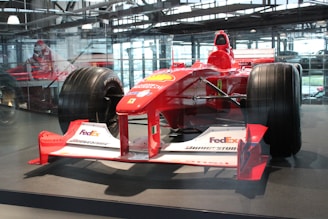 A red Formula 1 race car displayed in an indoor setting, featuring logos from various sponsors like FedEx, Bridgestone, and Magneti Marelli. The car is surrounded by a glass enclosure, with natural light filtering through the large windows in the background. The sleek design and large tires of the vehicle are prominently visible.