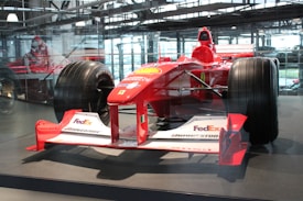 A red Formula 1 race car displayed in an indoor setting, featuring logos from various sponsors like FedEx, Bridgestone, and Magneti Marelli. The car is surrounded by a glass enclosure, with natural light filtering through the large windows in the background. The sleek design and large tires of the vehicle are prominently visible.