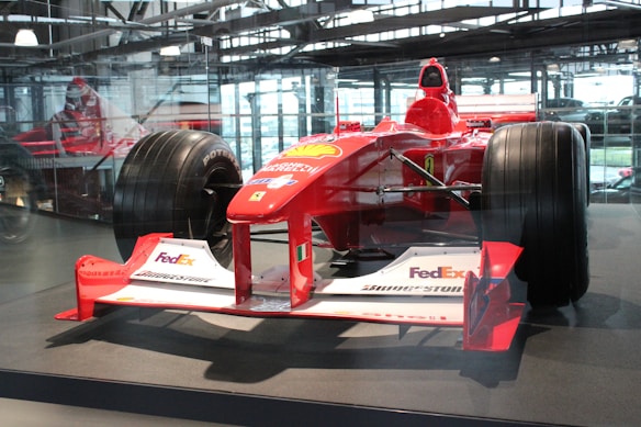 A red Formula 1 race car displayed in an indoor setting, featuring logos from various sponsors like FedEx, Bridgestone, and Magneti Marelli. The car is surrounded by a glass enclosure, with natural light filtering through the large windows in the background. The sleek design and large tires of the vehicle are prominently visible.