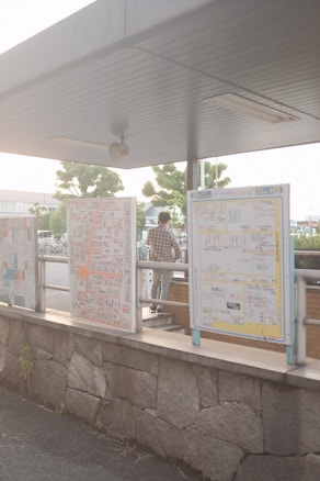A public transportation area with informational signs. The signs display detailed maps and information, likely in a bus or train station setting. The structure overhead provides shade, and a person is standing nearby, looking at the signs. Trees and a parking area are visible in the background.
