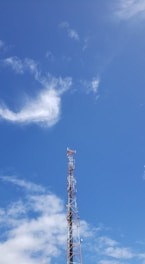 A telecommunications tower rises against a clear blue sky with scattered white clouds. The tower is equipped with various antennas and is composed of metal lattice supports.