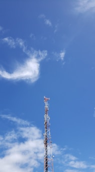 A telecommunications tower rises against a clear blue sky with scattered white clouds. The tower is equipped with various antennas and is composed of metal lattice supports.