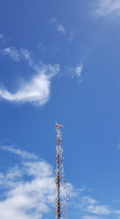 A telecommunications tower rises against a clear blue sky with scattered white clouds. The tower is equipped with various antennas and is composed of metal lattice supports.