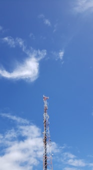 A telecommunications tower rises against a clear blue sky with scattered white clouds. The tower is equipped with various antennas and is composed of metal lattice supports.
