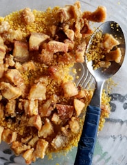 A close-up view of crispy fried pork belly pieces placed on a bed of yellow cornmeal. A metal spoon with a blue handle is partially submerged within the mixture.