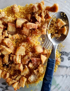 A close-up view of crispy fried pork belly pieces placed on a bed of yellow cornmeal. A metal spoon with a blue handle is partially submerged within the mixture.