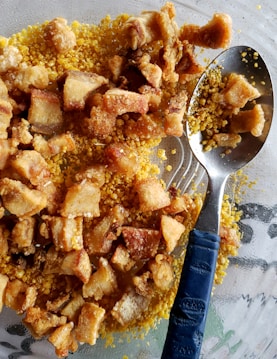 A close-up view of crispy fried pork belly pieces placed on a bed of yellow cornmeal. A metal spoon with a blue handle is partially submerged within the mixture.