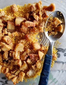 A close-up view of crispy fried pork belly pieces placed on a bed of yellow cornmeal. A metal spoon with a blue handle is partially submerged within the mixture.