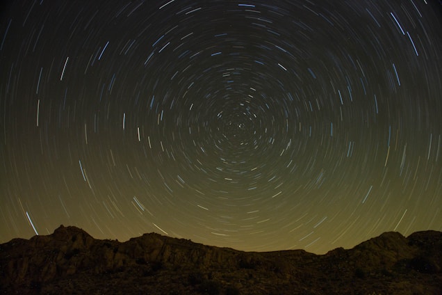 A long-exposure photograph capturing star trails in a circular pattern above a mountainous landscape. The night sky is filled with thousands of stars, creating a mesmerising spiral effect. The horizon shows rugged terrain with silhouetted peaks under the night sky.