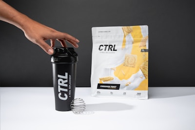 A hand reaches for a black shaker cup with the brand name 'CTRL' printed in white. Next to it, a package labeled 'Cinnamon Toast' displays images of cereal pieces with a splash of milk. The background is neutral gray.