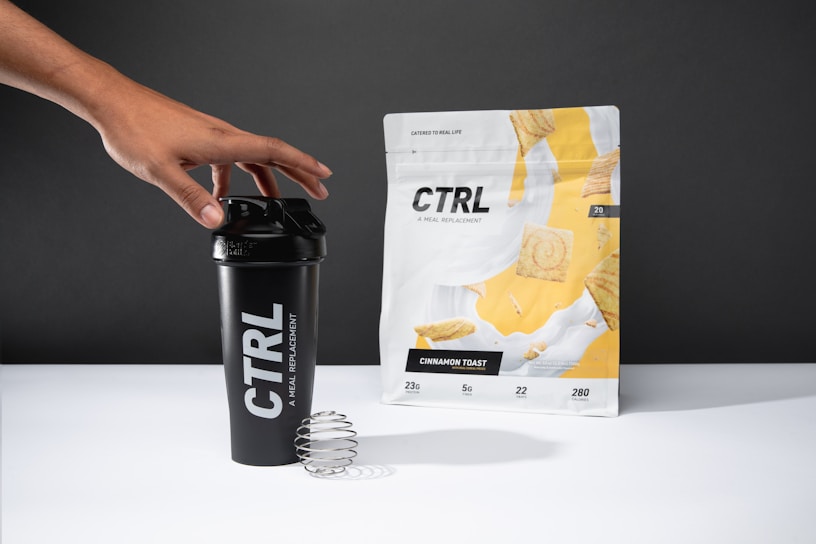 A hand reaches for a black shaker cup with the brand name 'CTRL' printed in white. Next to it, a package labeled 'Cinnamon Toast' displays images of cereal pieces with a splash of milk. The background is neutral gray.