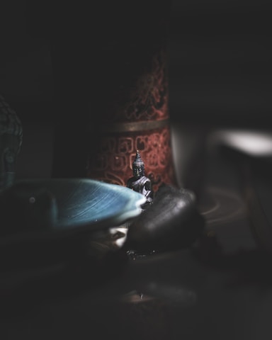 A small Buddha statue is placed on a reflective surface surrounded by dim, ambient lighting. The background includes an intricately designed object with reddish and green hues. A blue, leaf-patterned ceramic dish is partially visible in the foreground, contributing to the tranquil setting.