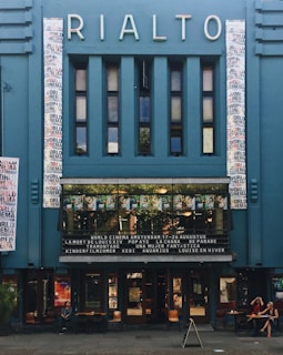 A vintage cinema facade with a blue exterior and the name 'Rialto' in illuminated letters. The facade includes a series of vertical rectangular windows and two vertical panels with multicolored text. Below, a marquee displays film titles. The entrance is lined with movie posters and two people are seated at a table on the sidewalk.