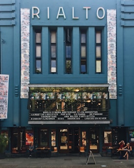 A vintage cinema facade with a blue exterior and the name 'Rialto' in illuminated letters. The facade includes a series of vertical rectangular windows and two vertical panels with multicolored text. Below, a marquee displays film titles. The entrance is lined with movie posters and two people are seated at a table on the sidewalk.