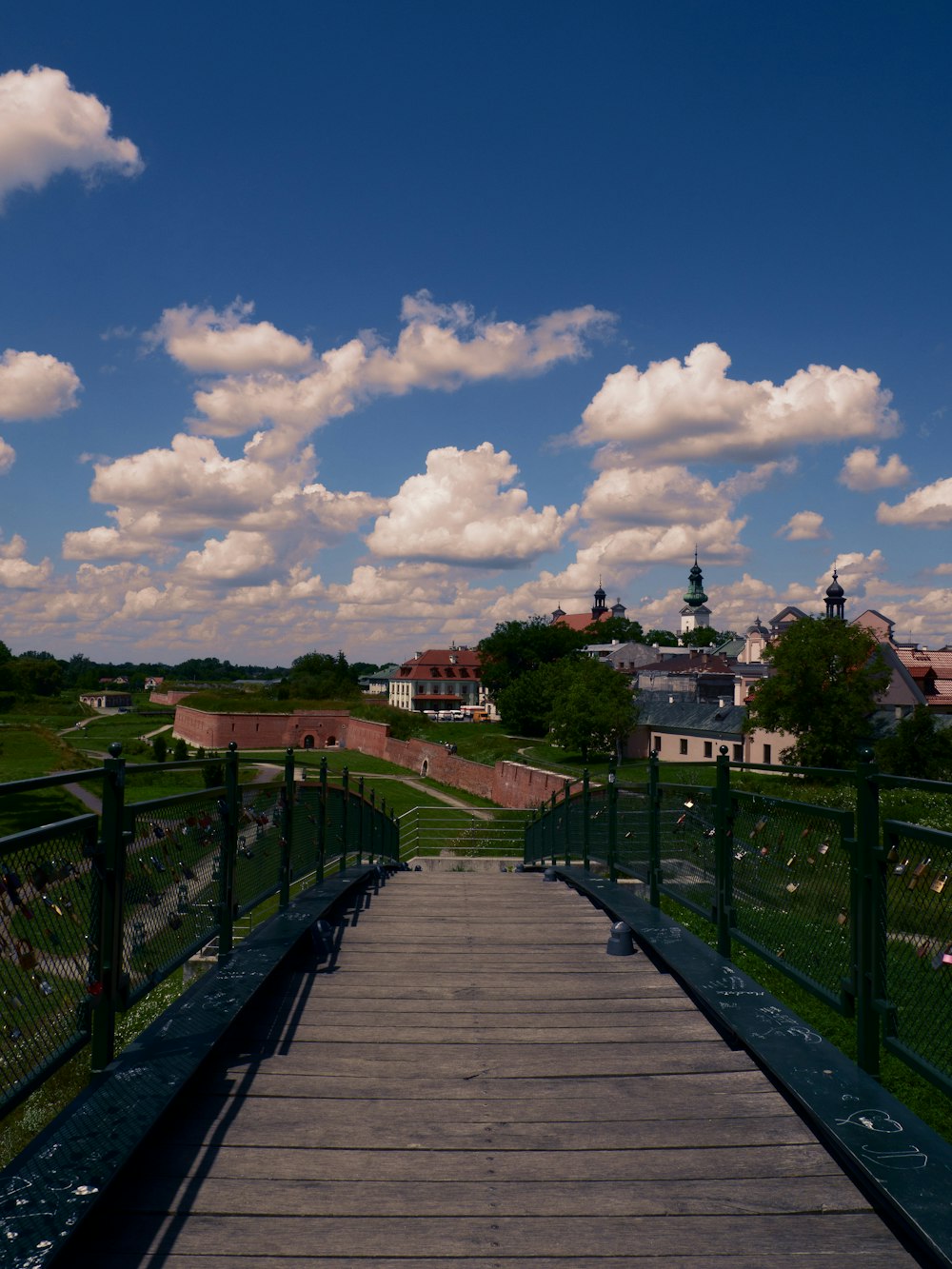 brown wooden bridge over green grass field under blue sky and white clouds during daytime