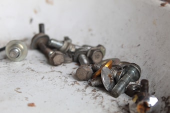 A collection of metallic bolts and screws scattered on a slightly dusty surface. The items vary in size and feature a mix of rust and shine, indicating some are older and worn while others are newer.
