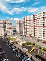 A residential area featuring several mid-rise apartment buildings with red and white facades. Cars are parked in designated parking spaces, and a small playground with a yellow slide is visible. The sky is bright blue with scattered clouds, indicating a clear, sunny day.