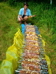 A person squats next to an array of skewers containing pieces of marinated meat, arranged on a large yellow sheet laid out on grass. The person holds one of the skewers, appearing to handle or inspect it.