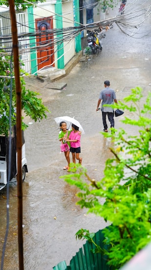 A street scene during a rain shower features two people sheltering under a single white umbrella. They appear to be smiling or laughing and are wearing casual clothes. Nearby, another person walks away, holding a plastic bag. The ground is wet with rainfall, and there are visible puddles. Surrounding the scene, lush green leaves are prominent, suggesting a tropical environment, and there is a red door in the background.