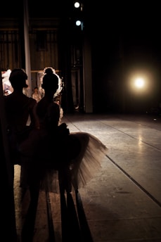 Two ballerinas are standing in the shadows backstage, highlighted by a bright light source that creates a dramatic silhouette effect. The stage floor is visible, and the atmosphere suggests anticipation.