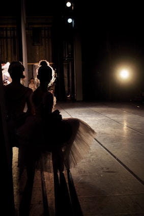 Two ballerinas are standing in the shadows backstage, highlighted by a bright light source that creates a dramatic silhouette effect. The stage floor is visible, and the atmosphere suggests anticipation.
