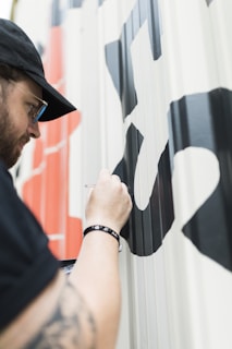 A person wearing a black cap and glasses is painting large black and red letters on a light-colored wall. They are holding a brush close to the surface as they apply paint. The focus is on the detailed work being done on the lettering.