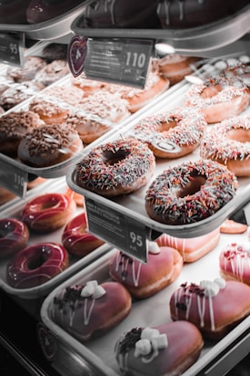 A display case filled with a variety of donuts, each with distinct toppings and glazes. Some donuts are covered with chocolate and colorful sprinkles, while others are topped with nuts, marshmallows, or a glossy fruit glaze. Price tags are visible on some trays.