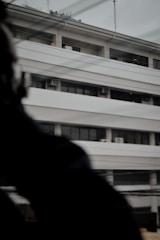 A multi-story building with a modern architectural design, featuring horizontal lines and balconies. The building has several air conditioning units visible on the exterior. There are some overhead wires and a blurred dark object or silhouette in the foreground, partially obscuring part of the building.