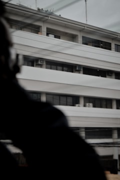 A multi-story building with a modern architectural design, featuring horizontal lines and balconies. The building has several air conditioning units visible on the exterior. There are some overhead wires and a blurred dark object or silhouette in the foreground, partially obscuring part of the building.