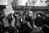 A group of individuals stand together, some holding a sign with the text '#Black Lives Matter.' One person is prominently raising a hand while speaking into a microphone. The crowd is diverse, with some wearing hoodies and masks. The setting appears to be an outdoor urban location with trees visible in the background.