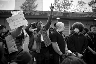 A group of individuals stand together, some holding a sign with the text '#Black Lives Matter.' One person is prominently raising a hand while speaking into a microphone. The crowd is diverse, with some wearing hoodies and masks. The setting appears to be an outdoor urban location with trees visible in the background.