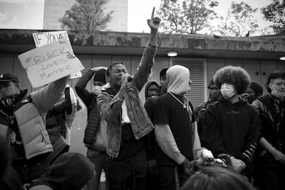 A group of individuals stand together, some holding a sign with the text '#Black Lives Matter.' One person is prominently raising a hand while speaking into a microphone. The crowd is diverse, with some wearing hoodies and masks. The setting appears to be an outdoor urban location with trees visible in the background.