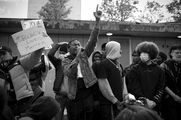 A group of individuals stand together, some holding a sign with the text '#Black Lives Matter.' One person is prominently raising a hand while speaking into a microphone. The crowd is diverse, with some wearing hoodies and masks. The setting appears to be an outdoor urban location with trees visible in the background.