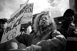 A group of people are participating in a protest with signs and a microphone. The central figure, a woman with curly hair, appears to be speaking or shouting. One of the signs reads 'DEFUND THE POLICE ACAB,' while another sign partially visible, contains text about neutrality and justice. The crowd is gathered outdoors under a cloudy sky.