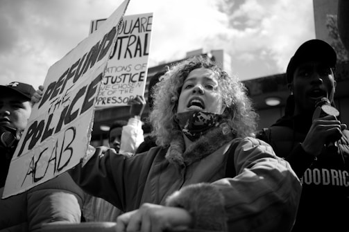 A group of people are participating in a protest with signs and a microphone. The central figure, a woman with curly hair, appears to be speaking or shouting. One of the signs reads 'DEFUND THE POLICE ACAB,' while another sign partially visible, contains text about neutrality and justice. The crowd is gathered outdoors under a cloudy sky.