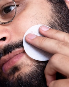 A person wearing glasses cleans their facial skin with a pad, focusing on the cheek area. The face has a beard and some facial hair.