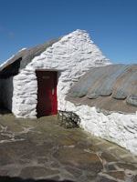 A whitewashed stone cottage with a thatched roof, featuring a bright red door. Fishing nets and equipment are placed outside the building, and the ground is covered with stone pavement. The sky is clear and blue, adding to the rustic charm of the scene.