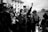 A group of people gathers in a public space, with one individual holding a microphone and raising a fist. Others are holding a megaphone and wearing masks. The scene takes place in front of a historic building.