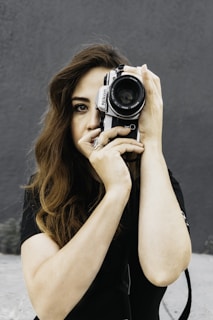 A person with long hair holding a Nikon camera up to their eye, seemingly about to take a photograph. The person has one eye visible, peering through the viewfinder. The background is a plain, dark gray wall, providing contrast to the image.