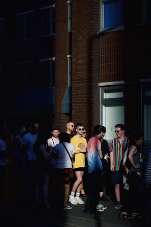 A group of people standing in a dimly lit urban area. Most individuals are wearing casual summer clothing, and some wear sunglasses. The background features a brick building with windows and a doorway, partially illuminated by sunlight.