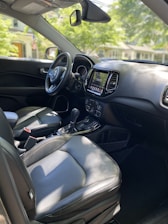 The image captures the interior of a modern car, showcasing the driver's seat, dashboard with a digital display, steering wheel, and gear shift. The materials appear to be a combination of leather and soft-touch surfaces. Sunlight filters through the windows, casting shadows and highlighting the details of the design.