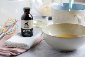 A bottle of organic vanilla extract is placed on a white cloth beside a colorful whisk and measuring cup. Two bowls are also seen, one appears to be mixing ingredients. The setting suggests a baking or culinary preparation scene.