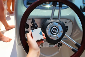 A hand is holding a dropper bottle labeled 'Batch Dream 1000 mg CBD' in front of a boat steering wheel. The dashboard of the boat is visible with various controls and gauges. There are barefoot individuals standing nearby, suggesting a relaxed setting.
