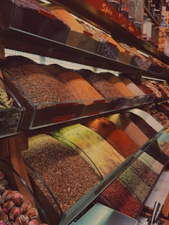 A vibrant and neatly organized display of various spices housed in transparent containers. The spices are arranged in rows and showcase a wide range of colors and textures, including shades of red, orange, green, and brown. Some spices are finely ground, while others are coarser or whole. The setting suggests a market or specialty spice shop.