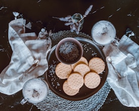A neatly arranged flat lay featuring a set of round biscuits on a dark plate, accompanied by a small bowl of dried lavender. Two glasses of frothy milk or coffee, garnished with lavender, are placed beside the plate. A woven placemat and two checkered napkins with silver spoons add texture to the composition. The scene radiates a rustic and cozy ambiance.