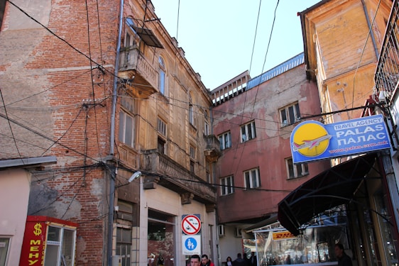 A narrow urban alleyway surrounded by old, weathered buildings with exposed brick and chipped plaster. A multitude of overhead electrical cables crisscross the scene. Visible signage includes a travel agency sign with a plane icon and Cyrillic text.