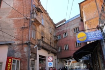 A narrow urban alleyway surrounded by old, weathered buildings with exposed brick and chipped plaster. A multitude of overhead electrical cables crisscross the scene. Visible signage includes a travel agency sign with a plane icon and Cyrillic text.