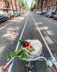 A bicycle is being ridden down a tree-lined urban street, filled with parked cars on either side. In the front basket of the bicycle, a reusable bag is filled with fresh produce including leafy greens, a zucchini, a red apple, and other fruits and vegetables.