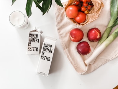 A collection of fresh produce and boxed water on a white surface. There are two cartons of Boxed Water, a candle, and a variety of fruits and vegetables including tomatoes, nectarines, leeks, and a bunch of cherry tomatoes wrapped in brown paper. Green leaves from a plant extend into the upper left corner.