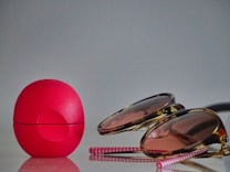 A bright pink spherical lip balm container sits on a reflective surface next to a pair of sunglasses with pinkish lenses and a gold frame. Two pink and white striped straws lie adjacent to these items.
